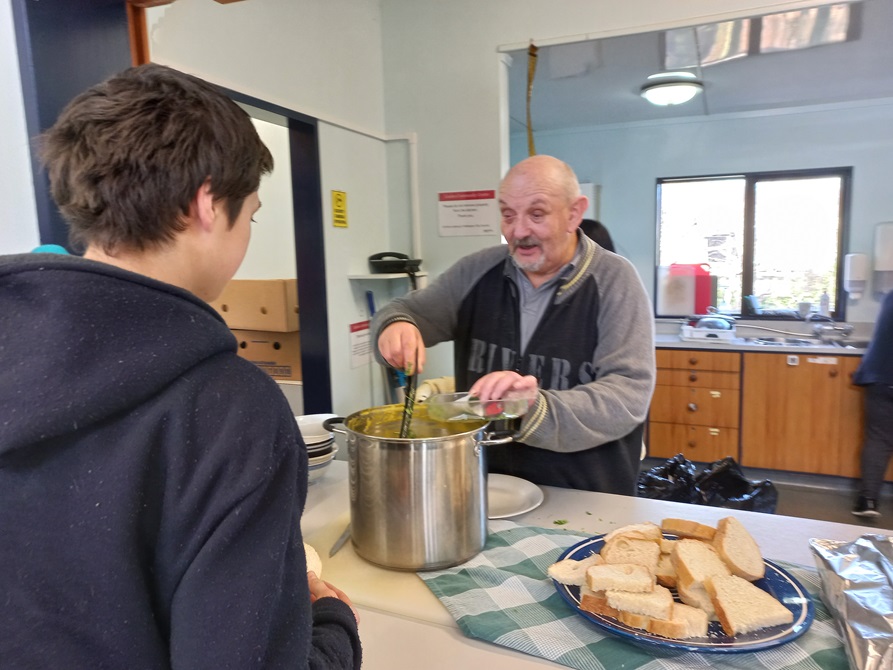 A young man standing at a counter where an older man, Colin, is stirring a large pot of soup, beside a plate of white sliced bread.