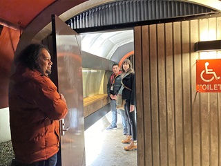 A man and a woman standing in a brightly-lit, spacious public toilet cubicle, with a man in an orange jacket holding the door open.