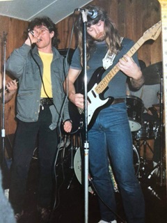 A low-angle shot looking up at two young men with long hair on stage performing, with the man on the left wearing a yellow t-shirt and grey jacket holding a microphone singing, and the man on right in blue jeans and grey t-shirt playing electric guitar.