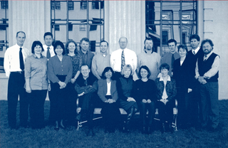 Thirteen men in ties and women standing in a row and five sitting in front on a bench posing in a black and white photograph for work, outside a building with white corrugated concrete façade and large reflective glass windows.