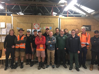 Fourteen people dressed in work boots and three in high-vis orange vests, standing grouped together, forward facing with arms folded, in a workshop with Fluro lights above.