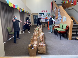 Three people wearing masks standing spaced apart with their arms spread in celebration, in a room with colourful bunting and dozens of paper bags filled with groceries lined up in the middle on the carpet.