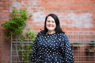 A smiling woman with mid-length black hair and a navy blue blouse with small white poker dots on it, standing in front of a blurred brick wall with plants growing up against it behind a metal frame.