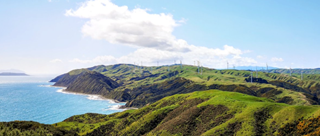 A blue ocean to the left, blue sky with white fluffy cloud above, and a vast green hilly landscape dotted with windmills into the distance.