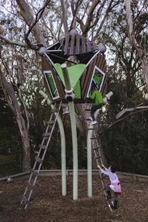 A new green and brown octagon-shaped tree house located with a very tall gum tree towering over it, situated on metal polls and with a ladder on each side with two children climbing up the ladder on the right side. 