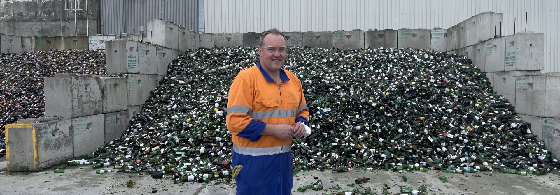 A man wearing overalls with bright blue pants and orange top, standing in front of a large pile of glass bottles sitting about a metre high, in-between a wall made of cinderblocks that's about five metres wide.