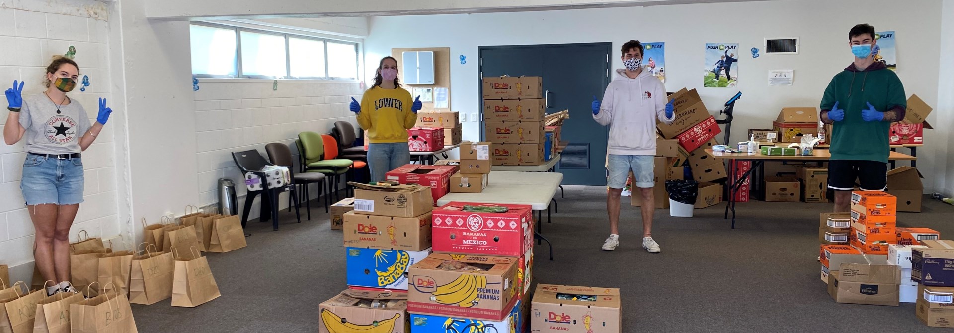 Four people wearing masks and spaced out across a large while and blue-carpeted room, standing in among dozens of paper bags and banana boxes while giving the thumbs up.