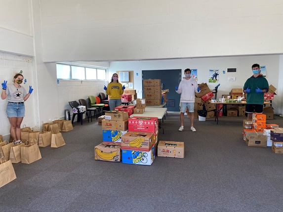 Four people wearing masks and spaced out across a large while and blue-carpeted room, standing in among dozens of paper bags and banana boxes while giving the thumbs up.