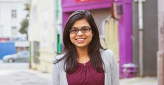 A smiling young woman with long black hair and glasses, wearing a maroon top and grey cardigan, standing in a laneway with grey, purple, and yellow painted buildings.