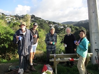 Four men and a young woman all dressed in warm outdoor gear, standing on a bushy hill next to a park bench, all holding a cuppa, with shovels and tools on the ground.