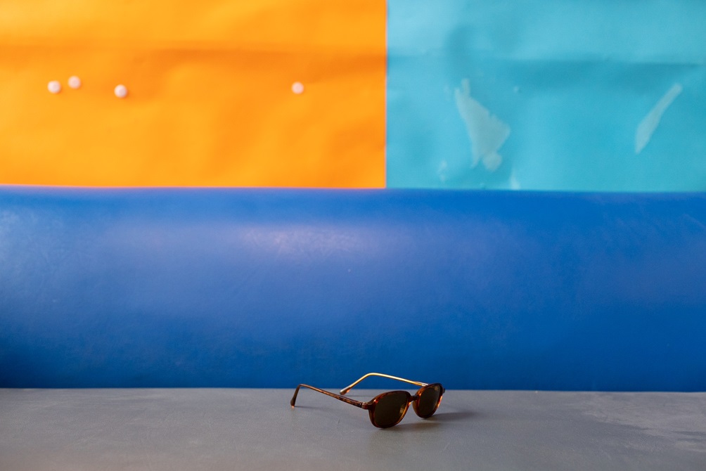 A pair of brown sunglasses abandoned on a blue and grey couch with colourful squares hanging on the wall in the background.