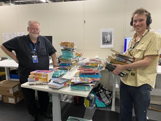 A smiling solid-framed gentleman on the left in dark shirt and pants with is hand on his hip, standing next to a table piled with books, and a younger lad to the right with headphones on and a tan shirt holding a stack of books.