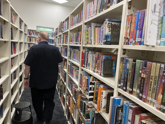 The back of a large-framed man with dark coloured clothing walking through a library isle of shelving filled with books on either side.