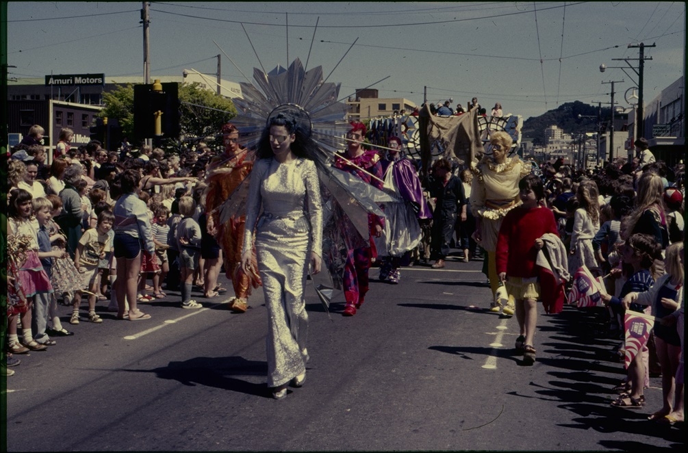 Men and women in fantastical metalic and shiny costumes walk in a street parade as families watch from the sides.