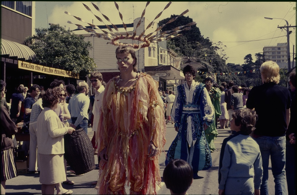 Men dressed in colourful, draping garments and large headdresses walk down a street watched by a crowd of people.