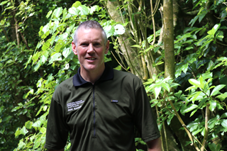 Mark Kent with short hair wearing his green ranger uniform shirt standing in front of green trees and bush.