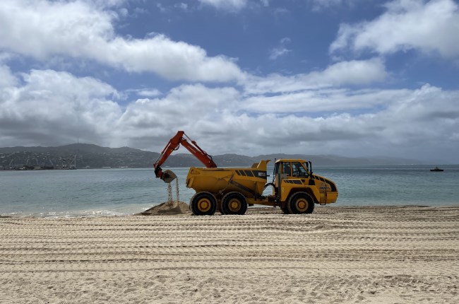 A yellow digger truck scooping up sand on the beach by the shoreline, with the water and land in distance behind and golden sand in foreground.