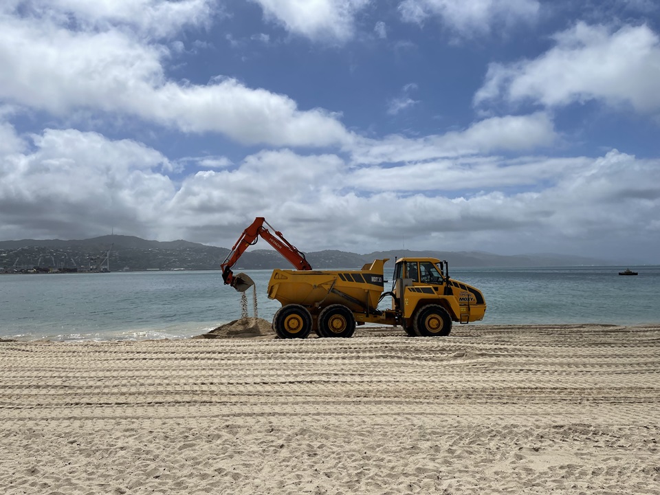 A yellow digger truck scooping up sand on the beach by the shoreline, with the water and land in distance behind and golden sand in foreground.