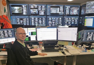 Tim Kirby sitting at a desk with a wall of screens behind him.