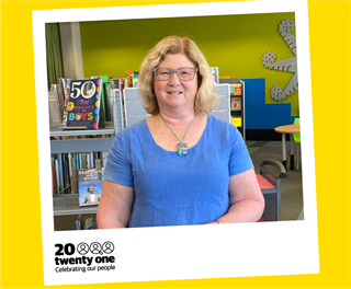 A photo Raewyn in a blue top standing in front of a library shelf full of books.