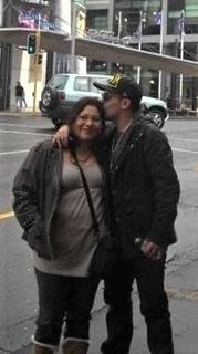 A man wearing cap and sunglasses with his arm around a woman, kissing her on her head, as they stand beside a road and buildings.