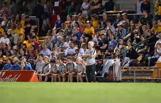 A line-up of soccer players sitting in the benches on the field with stadium seats packed with fans behind. 
