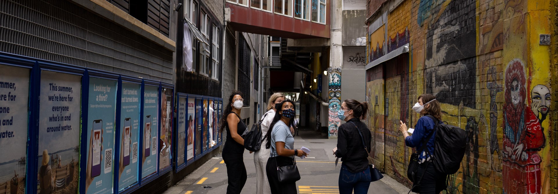 Five young woman, dressed casually and wearing masks, walking down an alleyway with posters on the left, a bright mural on the right, and a covered brown overbridge ahead.