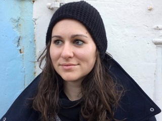 A young woman with dark long hair and a black jacket and beanie, looking left with a slight smile, in front of a blue and white wall on which the paint is beginning to chip.