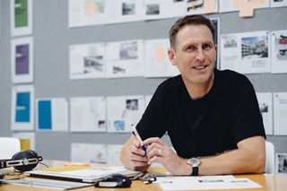 A man in a black t-shirt sitting at a wooden desk with paperwork on it and pinned to the wall behind him.