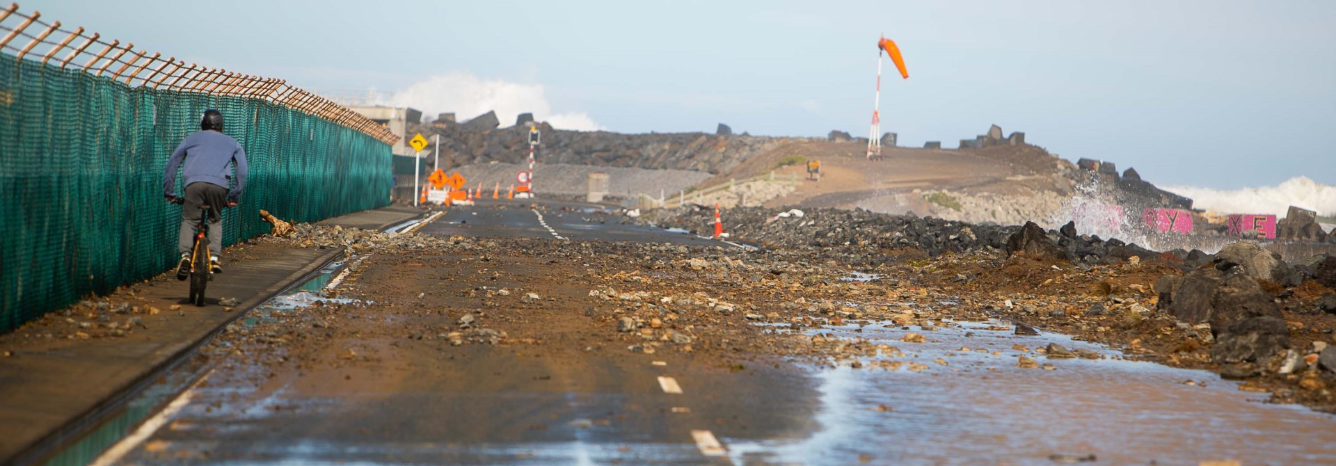 A cyclist riding over debris and rocks strewn over a wide road, which runs alongside the coast. The sky is blue following the storm.