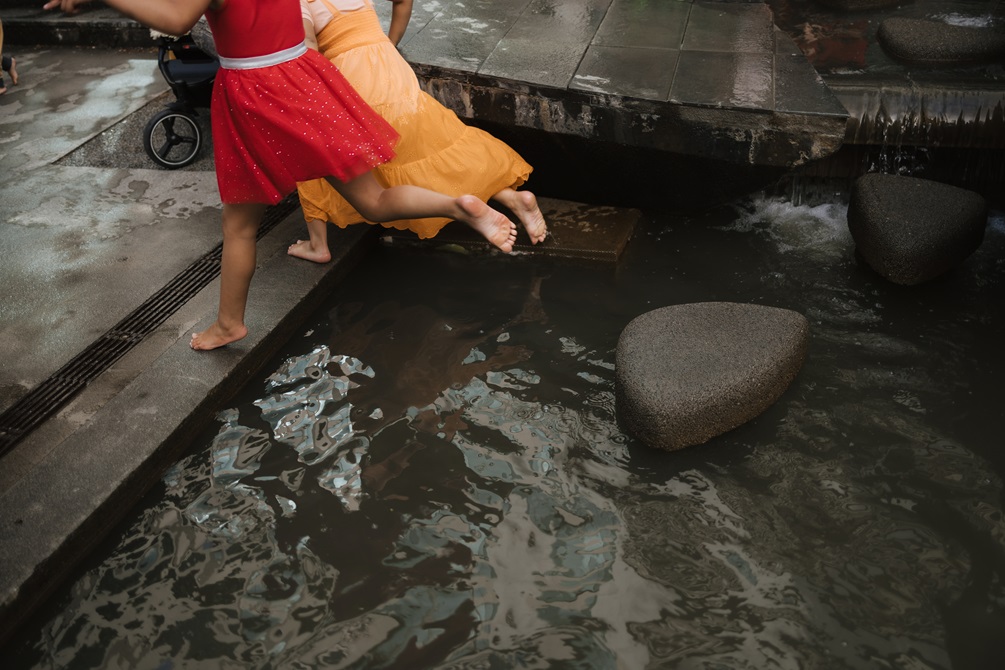 Two children wearing red and orange dresses jump over water in unison from a rock to a step. They are surrounded by wet concrete.