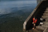 A young woman wearing a red cardigan and headphones, sitting with bare feet on concrete steps next to the ocean, as she reads a book with her belongings beside her.