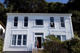 A two storey white house with a person leaning against the front door wearing a face mask, and another person leaning out a window on the second floor.