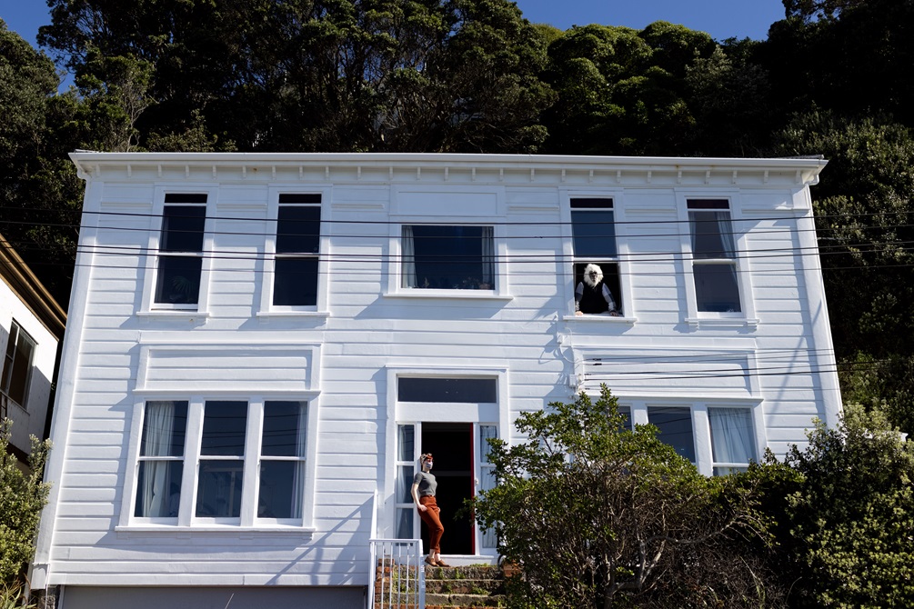 A two storey white house with a person leaning against the front door wearing a face mask, and another person leaning out a window on the second floor.