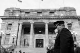 A man wearing a dark official uniform and a mask, standing in a row alongside other people outside a grand government building with a staircase and tall marble pillars at the entrance. 