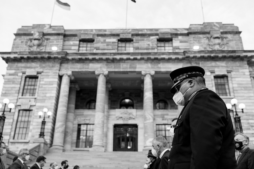 A man wearing a dark official uniform and a mask, standing in a row alongside other people outside a grand government building with a staircase and tall marble pillars at the entrance. 