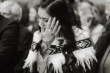 A young woman dressed in a special korowai cloak. Her hand, which is tattooed in Māori design, is brushing her dark hair from her face.