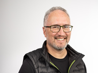 Portrait of Wellington City Council mana whenua representative for Taranaki Whānui ki te Upoko o te Ika, Holden Hohaia. He is smiling in front of a white wall, wearing glasses, and is dressed in a black tee shirt and puffer vest.