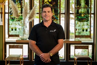 Man standing in a chapel smiling, wearing a black shirt.