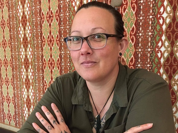 Person wearing a green shirt and glasses, sitting infront of a colourful patterned wall.