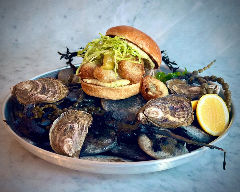 Half Doz burger by GPO on plate surrounded by oysters and seaweed.