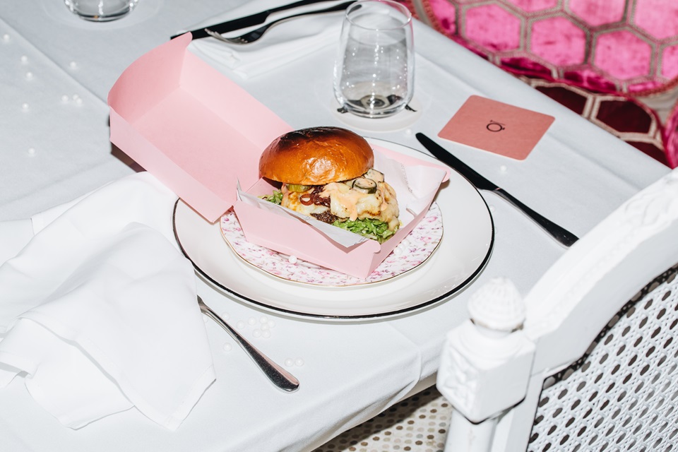 Hippopotamus Museum burger in a pink container surrounded by fine dining setting.