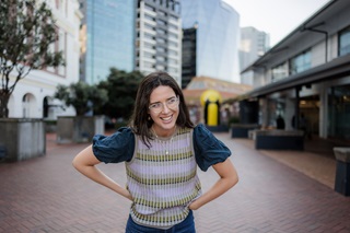 A woman wearing a colourful vest and a puffy sleeved navy top smiling with her hands on her hips.