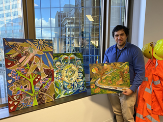 Team Leader Matai holding a painted canvas, with two other pieces of art sitting on the windowsill.