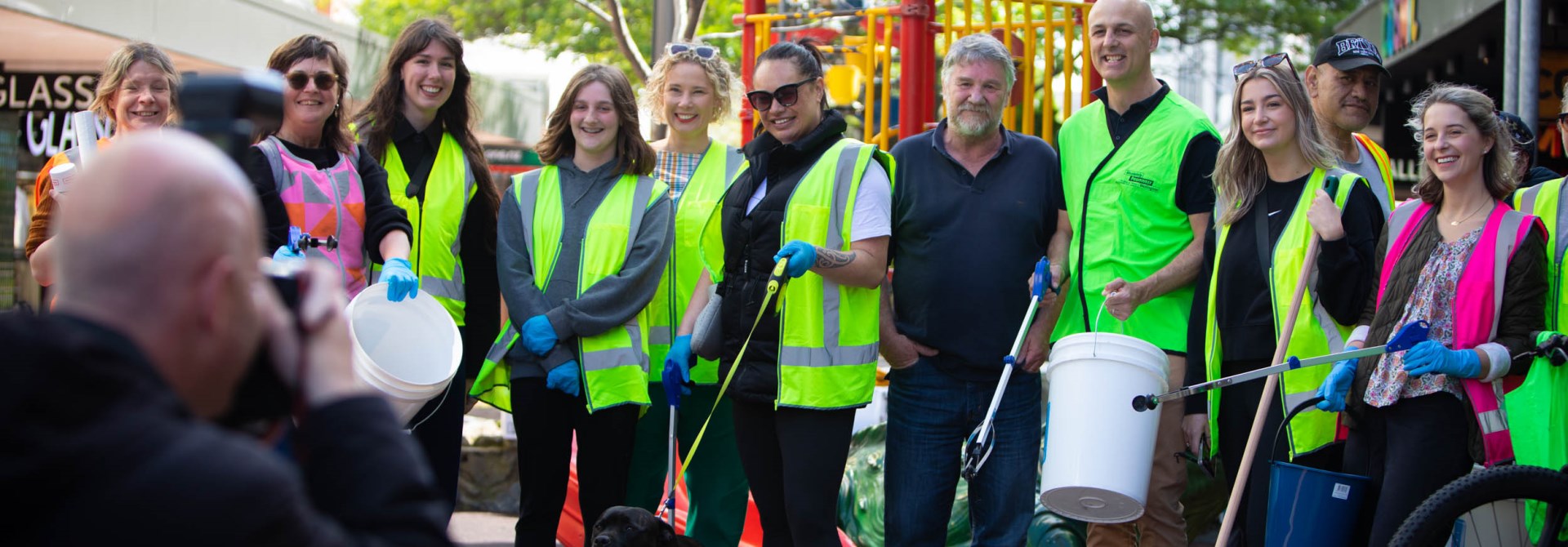 About a dozen people standing in a line wearing yellow high-vis vests and gloves, holding cleaning buckets and sticks for picking up rubbish.