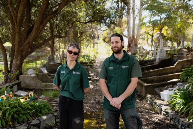 Taylor and Jayden stand in their green polo shirt uniforms under the trees in a cemetery.