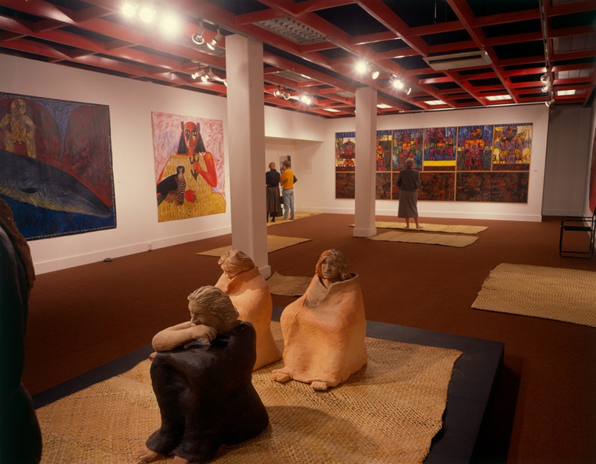 An exhibition at City Gallery Wellington featuring sculptures by Shona Davies of figures sitting on the floor and bright colourful paintings of Māori women by Robyn Kahukiwa.