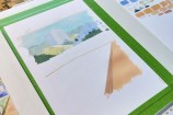 The beginnings of a painting on a desk, with colour swatches to the right and paint brushes on in jars.
