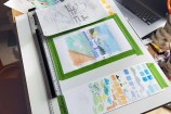 A desk with a dark grey cat, open laptop, and watercolour paintings on it.