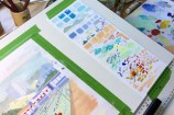 A watercolour painting of an outdoor pool on a desk, with colour swatches on the right and jars of paintbrushes and pencils.